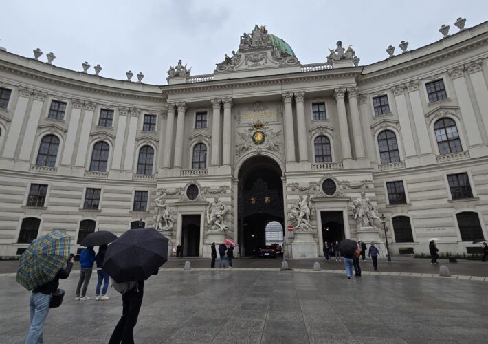 Grand, ornate historic building with tall columns and statues, people holding umbrellas on a rainy plaza in front of a central archway.