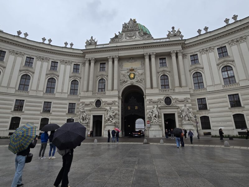 Grand, ornate historic building with tall columns and statues, people holding umbrellas on a rainy plaza in front of a central archway.