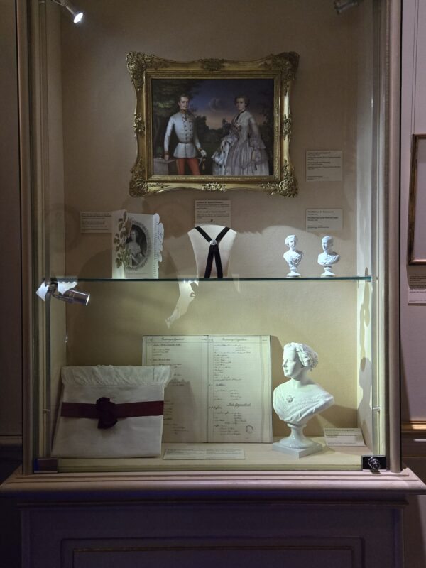 Ornate gold-framed portrait of a man and woman in historic dress, displayed in a museum case with busts and a cushion nearby
