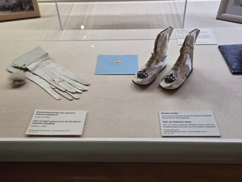 White vintage gloves and a white lace boot display with a light blue envelope in a museum case, with German labels nearby.