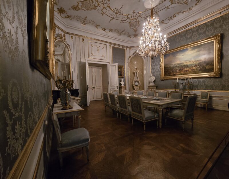 Elegant antique dining room with a long table, upholstered chairs, and a crystal chandelier in a gilded, ornate setting.