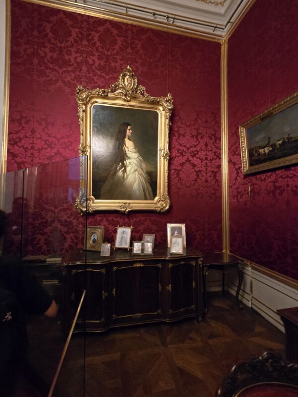 Ornate gold-framed portrait of a woman in a white gown on a red damask wall, with small framed photos on a dark desk below.