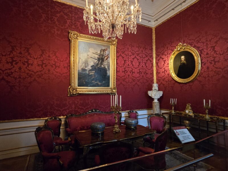 Opulent room with red damask walls, a crystal chandelier, and gold-framed paintings (ship scene and portrait) on the wall red by a formal parlor setting.