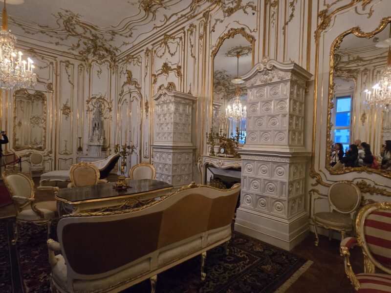 Luxurious rococo parlor with gold-edged white panels, mirrors, chandeliers, and antique seating.