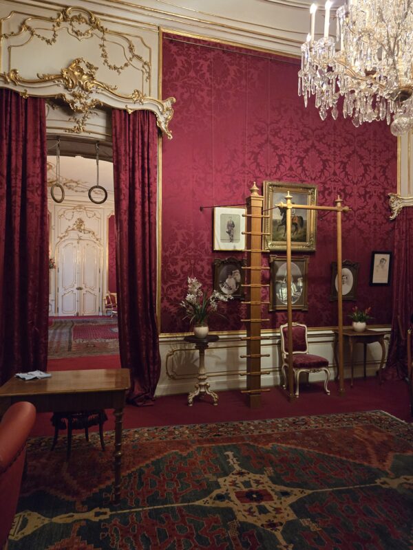 Ornate room with burgundy damask wallpaper, gold-trimmed archway, and crystal chandelier overhead.