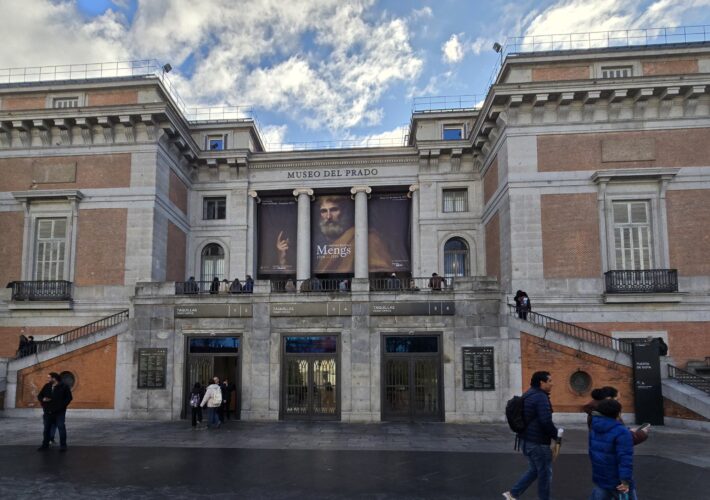 Facade of the Museo del Prado in Madrid with banners advertising a Mengs exhibit, stone and brick exterior, steps and visitors in front.