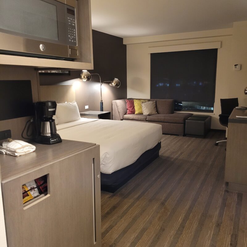 Hotel room with a bed in foreground, kitchenette counter, microwave, and a sofa with colorful cushions under a window with blinds.