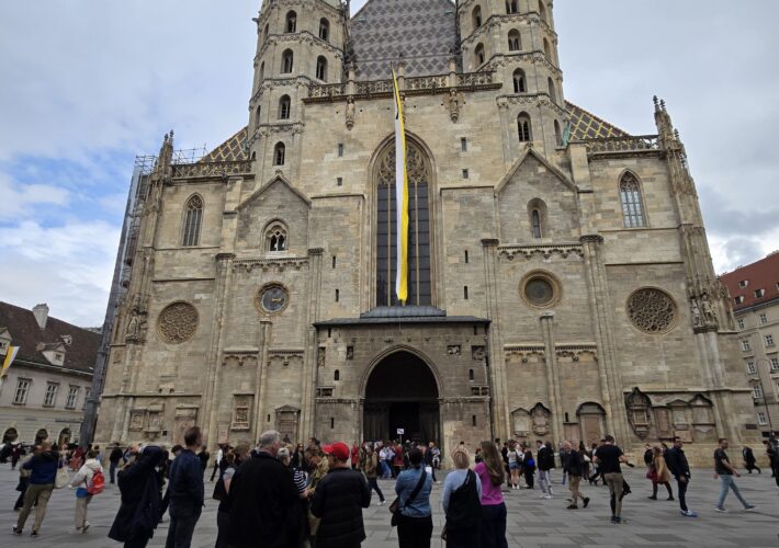 Front view of a large stone cathedral with two tall towers, Gothic windows, and a striped tile roof, people gathered in the foreground.