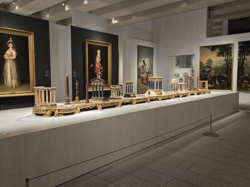 Museum gallery with tall ornate portraits in gold frames along dark and light walls, and a long display of intricate miniature architectural models on a raised white platform.