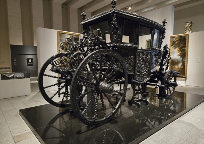 Ornate black carriage with intricate ironwork on display in a museum, surrounded by framed paintings on light walls.