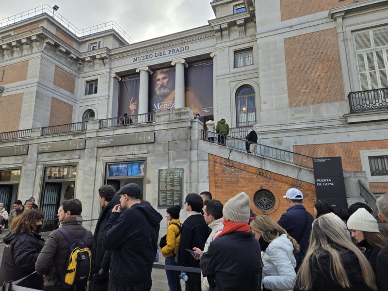 Crowd of visitors gathered outside the Prado Museum entrance, with ticket windows and large banner advertisements on the façade.