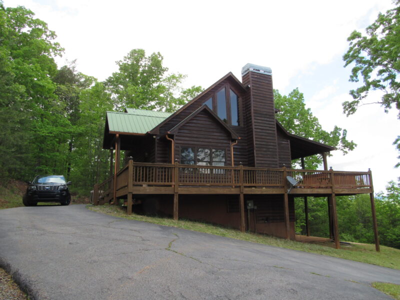 Two-story log cabin with green metal roof and large windows, set among green trees; a dark SUV is parked on the sloped driveway to the left.