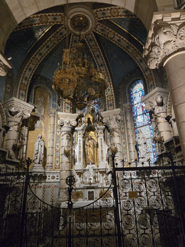 Ornate church interior with gilded altar, statues, and a decorative iron railing in front of detailed mosaic arches and stained glass.