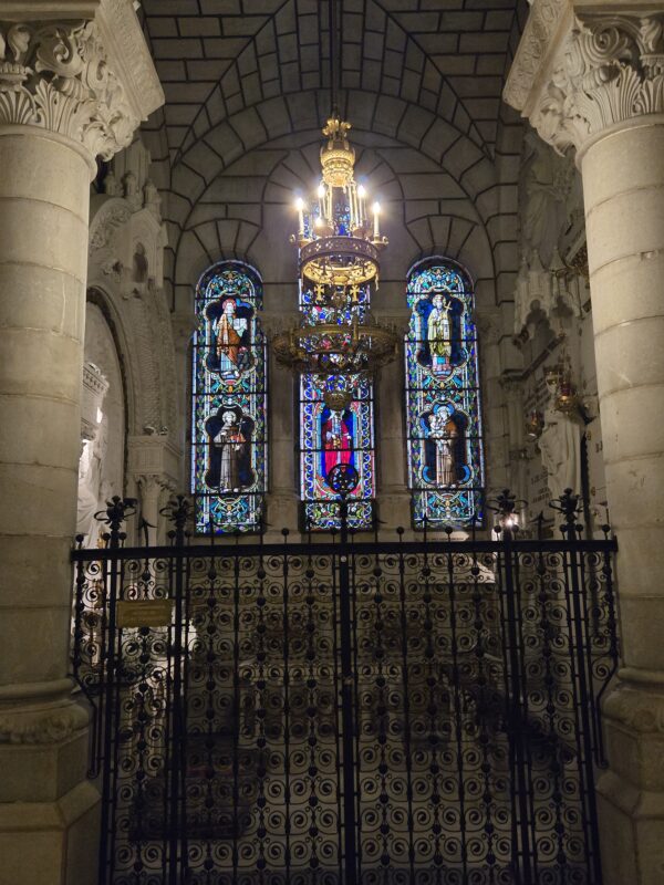 Interior of a church showing a gold chandelier hanging between three tall, colorful stained-glass windows, with carved stone columns on either side and a decorative wrought-iron railing in the foreground.