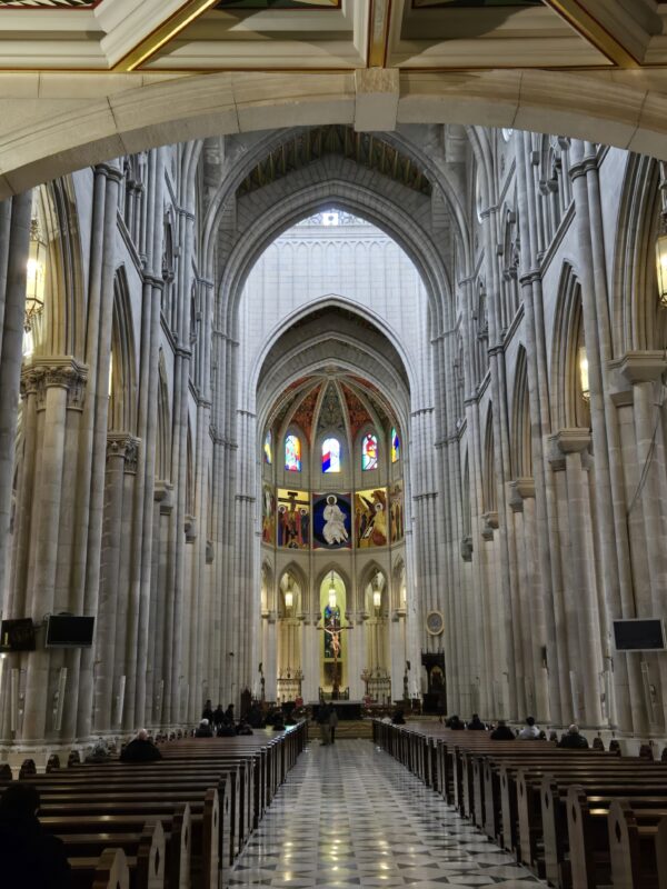 Interior of a Gothic cathedral nave with tall stone arches, rows of pews, and colorful stained-glass windows at the far end