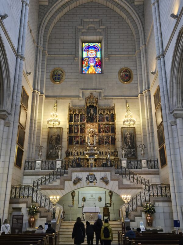 Interior of a grand church with an ornate altar, stained glass window, and arched stone walls. People sit and stand in the nave.