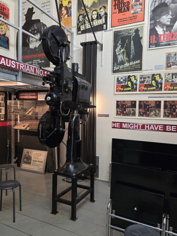 Vintage film projector on a stand in a museum-like room, surrounded by old movie posters on the wall.