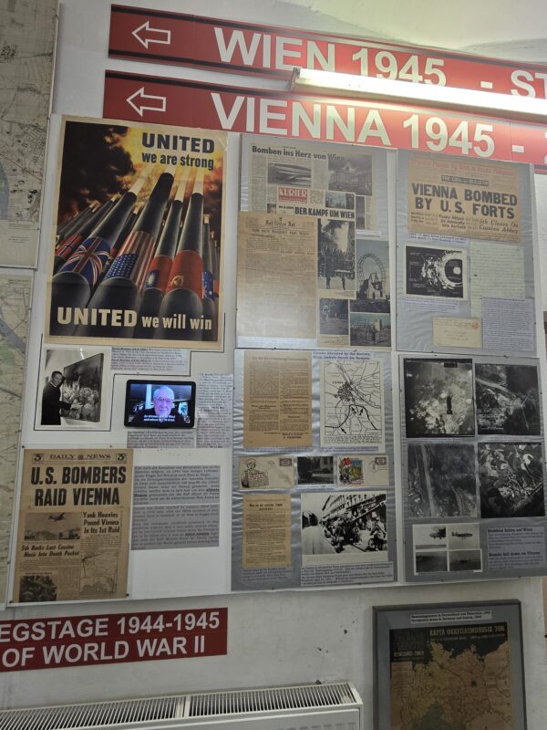 Wall display of World War II–era newspaper clippings and banners about Vienna 1945, including a 'UNITED we will win' poster and headlines about the bombing campaign nearby on the wall.