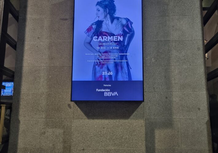 Vertical blue-lit theater poster advertising Carmen by Georges Bizet, featuring a woman in an off-shoulder dress against a blue background mounted on a stone wall.