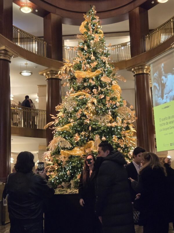 Large Christmas tree with gold ribbons and white lights in a grand lobby, with people gathered around taking photos.