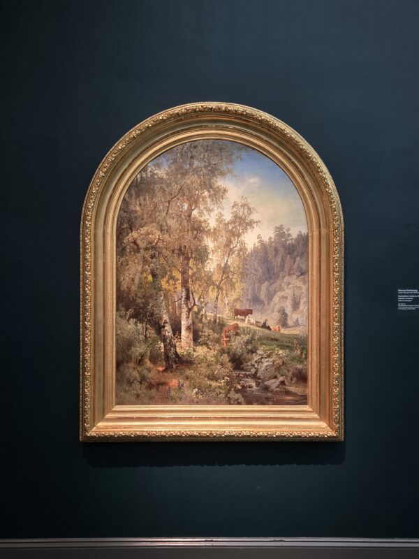 Landscape painting of a forest with trees and cows in a gold ornate frame mounted on a dark blue wall.