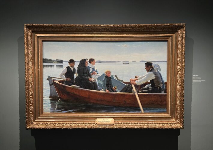 Group of people in a wooden rowboat on a calm lake, with a woman in a black shawl and a child at the bow, by the shore.