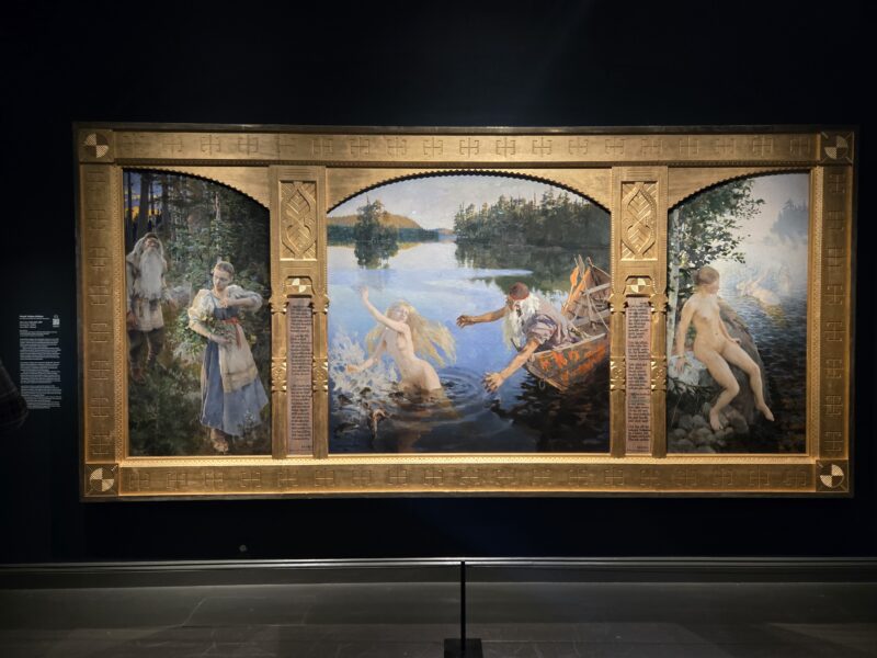 Large painting framed in an ornate gold frame, showing a forested lakeside scene with several figures, including nude ones by the water, on display in a gallery