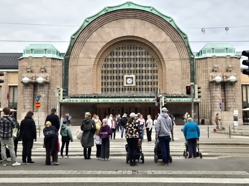Crowd of people at a busy city street in front of a grand arched train station facade with a clock above the entrance.