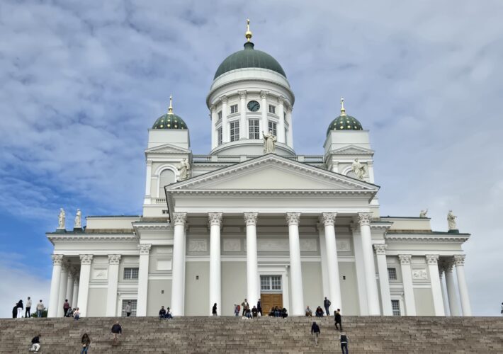 White neoclassical building with green domes, statues, and broad stairs populated by visitors.