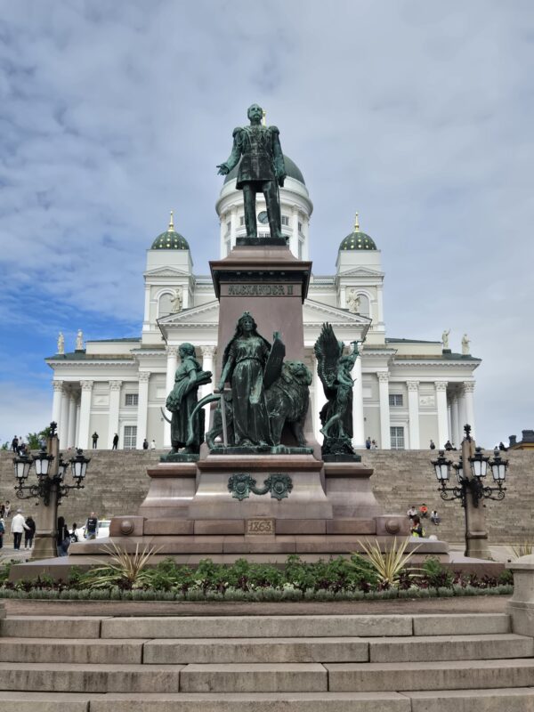 Bronze statue of Alexander II on a pedestal in front of a grand white domed building with green domes and columns behind.