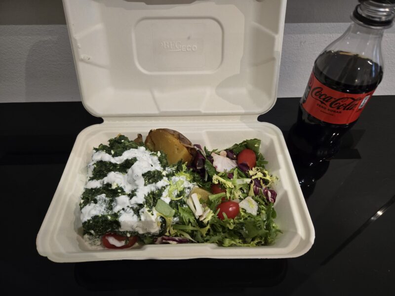 Takeout container filled with a mixed salad (greens, cherry tomatoes, feta with herb dressing) next to a bottle of Coca‑Cola Zero Sugar.
