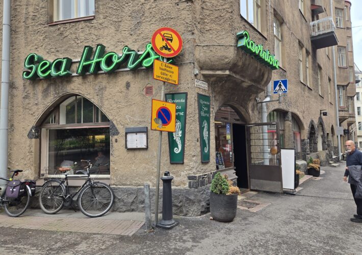 Rustic stucco building storefront with a bright green neon sign reading 'Sea Horses' and street signs, bicycles parked outside, and a pedestrian crossing sign nearby.