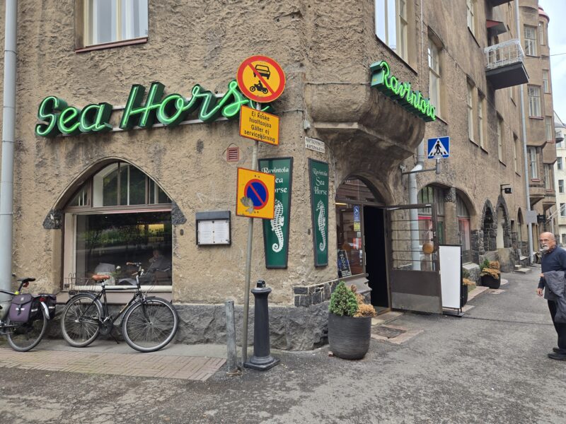 Rustic stucco building storefront with a bright green neon sign reading 'Sea Horses' and street signs, bicycles parked outside, and a pedestrian crossing sign nearby.