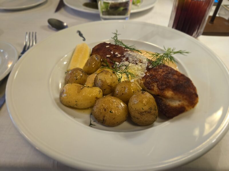 Plate of crispy breaded fish with roasted new potatoes, lemon wedge, and dill garnish on a white dish? text implies informativie—note: ensure concise phrasing.