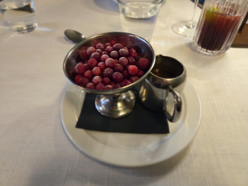 Close-up of a stainless steel bowl with frozen red berries dusted with sugar, resting on a saucer with a black napkin and blurred table setting in the background.