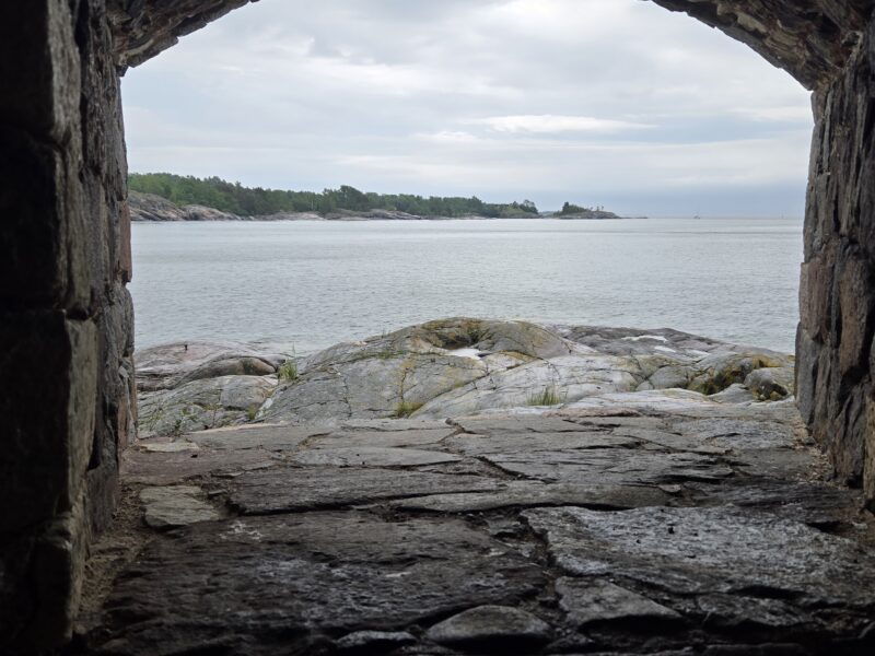 Rocky cave opening framing a calm sea with a tree-covered island in the distance under a cloudy sky.