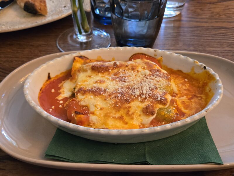 Ramekin with baked pasta (lasagna) covered in melted cheese and parmesan, served on a white plate with a green napkin on a wooden table.