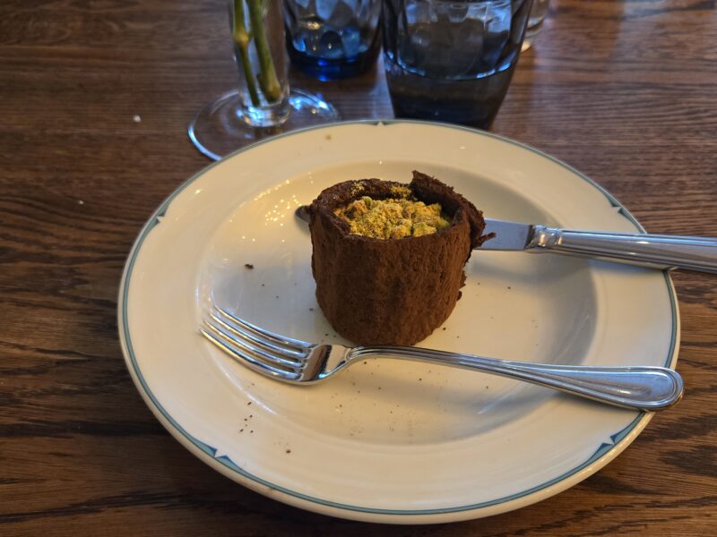 Chocolate tart on a white plate with teal rim, crumbs around it, butterknife and fork resting nearby on a wooden table. Behind are a glass and a dark beverage.