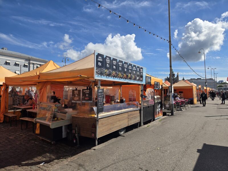 Row of bright orange market stalls and a long menu board under a clear blue sky with string lights above the street.