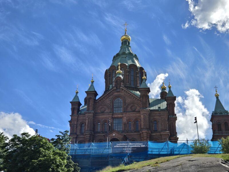 Ornate brick church with green copper domes and golden onion domes topped by crosses, set against a blue sky; construction fencing at the base.
