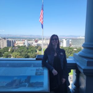 Smiling woman in a denim jacket and sunglasses standing on a rooftop with an American flag in the background and a city skyline beyond.