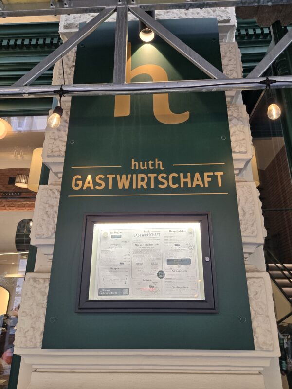 Green storefront sign reading 'huth GASTWIRTSCHAFT' with a lit display window showing a menu framed in black beneath, mounted on a stone façade with string lights nearby.