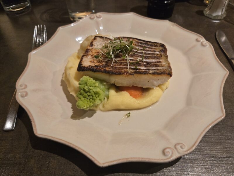 Grilled-seared white fish atop mashed potatoes with broccoli florets and microgreens garnish on a decorative plate.
