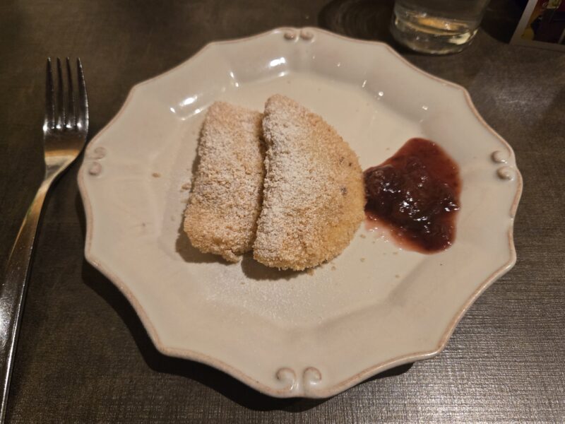 Two breaded fried sticks dusted with powdered sugar on a light plate, with a spoonful of red jam nearby on the right.