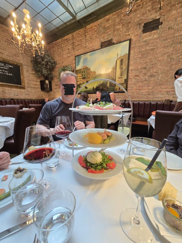 Man with blurred face seated at a white-tablecloth table in a brick-walled restaurant, with a two-tier seafood and herb platter in front.