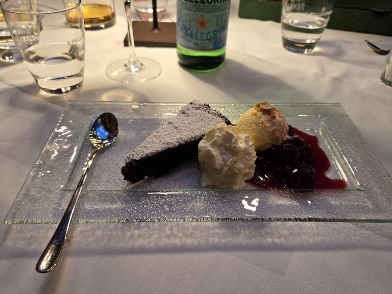 Dessert plate with chocolate cake dusted in sugar, vanilla ice cream, whipped cream, and berry sauce on a glass platter, with a spoon nearby.