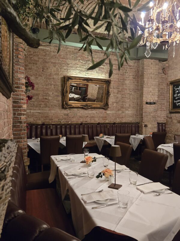Elegant brick-walled dining room with a gold-framed mirror, chandelier, and white-tablecloth tables set with glassware and orange roses.