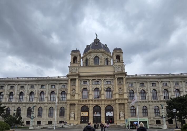 Facade of a grand historic museum with a central domed tower and two side towers under a cloudy sky, Vienna’s architecture style evident in stonework and arches