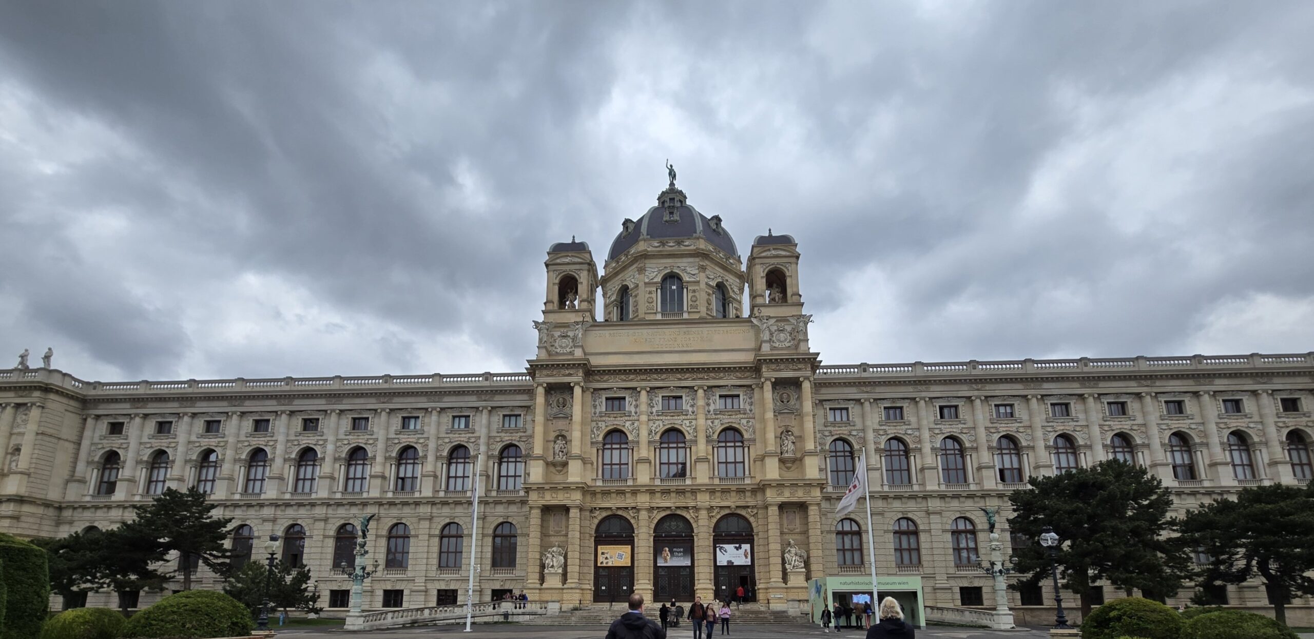 Facade of a grand historic museum with a central domed tower and two side towers under a cloudy sky, Vienna’s architecture style evident in stonework and arches