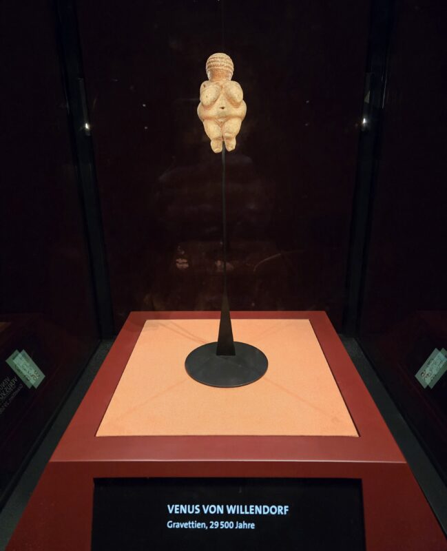 Venus von Willendorf figurine on a pedestal inside a glass display case, with a label reading 'VENUS VON WILLENDORF' and 'Gravettien, 29 500 Jahre'.